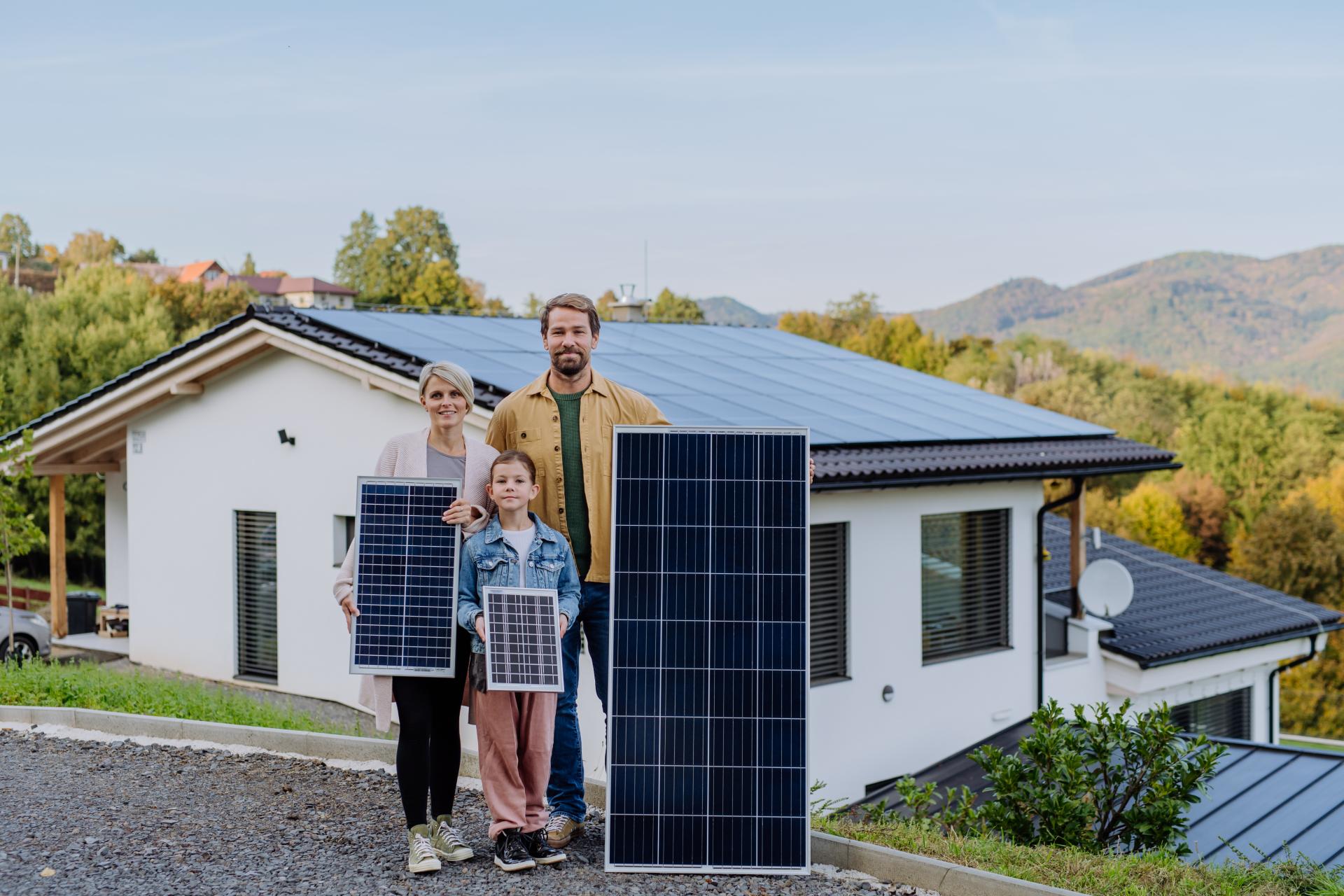 Rodina stojí před domem, drží fotovoltaické panely. Jak ušetřit za elektřinu s rostoucí rodinou?