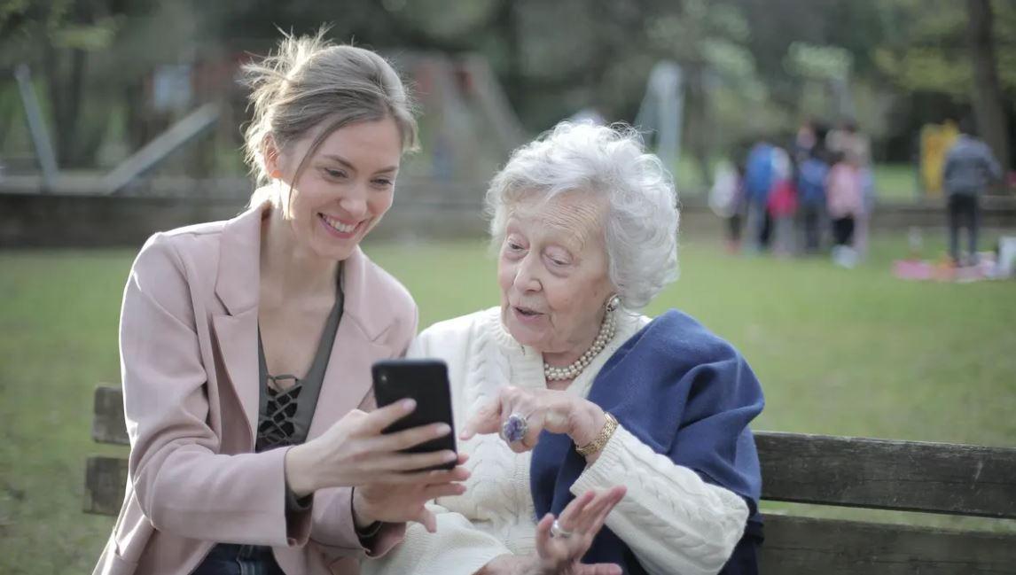 Mladá žena vysvětluje seniorce aplikaci v mobilu. Samota a pád? Díky tísňové péči přišla pomoc včas