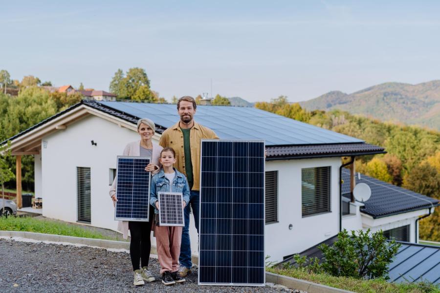 Rodina stojí před domem, drží fotovoltaické panely. Jak ušetřit za elektřinu s rostoucí rodinou?